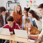 Estudantes colaboram em laptops em uma sala de aula, orientados por um professor.