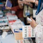 Livros expostos em uma feira literária enquanto visitantes circulam e observam os títulos.