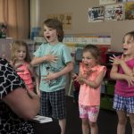 Crianças aprendem língua de sinais com uma professora em uma sala de aula colorida.