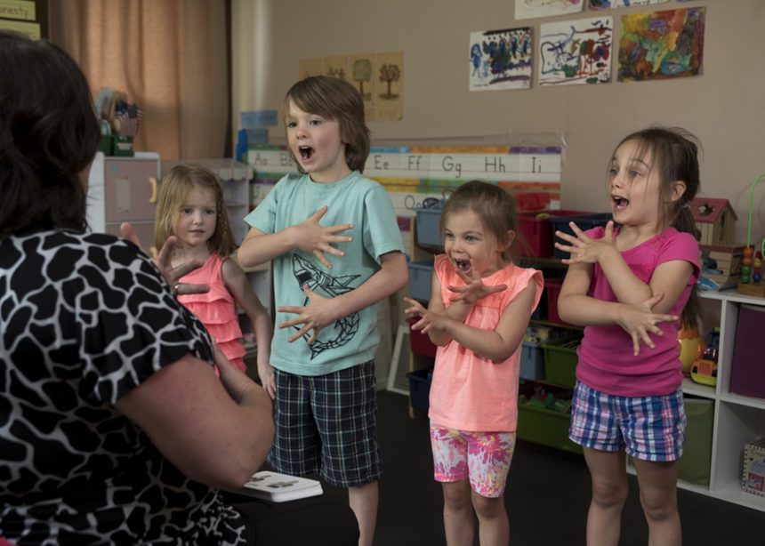 Crianças aprendem língua de sinais com uma professora em uma sala de aula colorida.