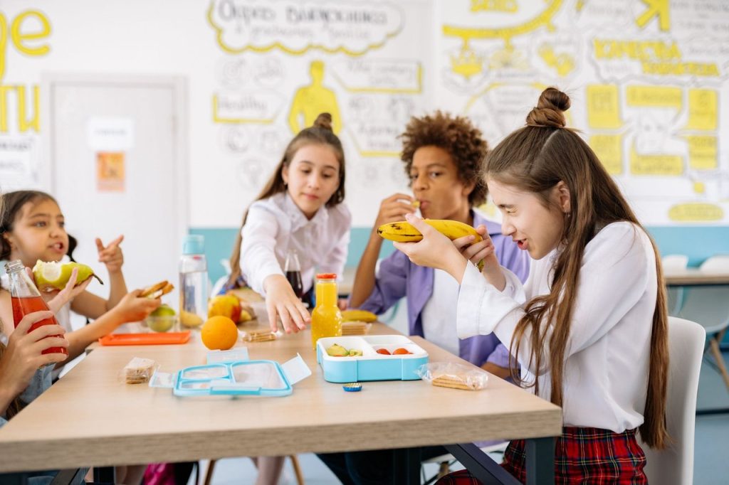 Estudantes compartilham lanches e bebidas em uma mesa de refeitório escolar.