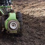 Agricultor opera um trator de pequeno porte em campo recém-arado para preparar o solo.